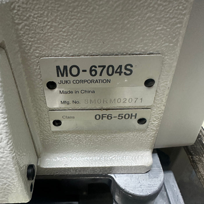 Close-up of a metal product label on a textured off-white machine housing, showing the model MO-6704S, JUKI CORPORATION, Made in China, and Class 0F6 50H: 1 Needle(3 Threads) Overlock | 4.8mm 3 rolls | Medium-Heavy Running Stitch(Denims) | Extra High Throw
