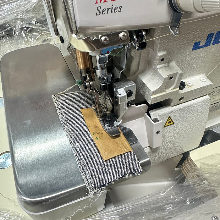 Close-up of a Juki industrial serger sewing machine in silver and white, actively sewing Denim. Juki MO-6700 Series overlock sewing machine. MO6704S 0F6 50H: 1 Needle(3 Threads) Overlock | 4.8mm 3 rolls | Medium-Heavy Running Stitch(Denims) | Extra High Throw