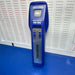 A blue handheld needle detector device labeled 'MACHINE DETECTOR HN-305' and 'HASHIMA' stands upright on a blue surface in front of a white sheet covered with repeated 