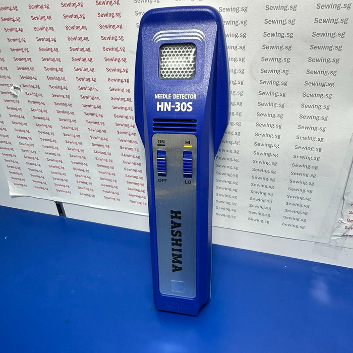 A blue handheld needle detector device labeled 'MACHINE DETECTOR HN-305' and 'HASHIMA' stands upright on a blue surface in front of a white sheet covered with repeated 'Sewing-sg' text.