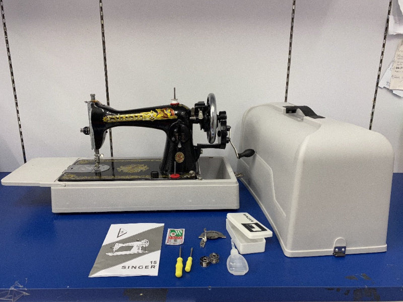 A traditional Singer sewing machine with a portable white box. The machine has a manual hand crank drive and is accompanied by various accessories such as bobbins, needles, and a needle plate.